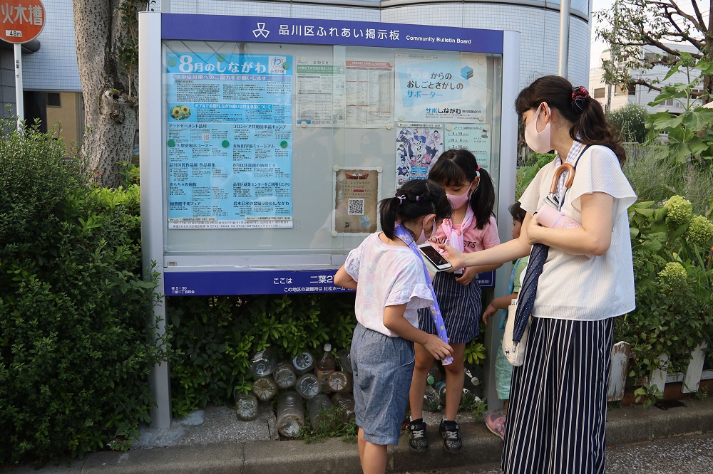 品川区ふれあい掲示板の前でえばこクエストを楽しむ親子、女の子3人と母親