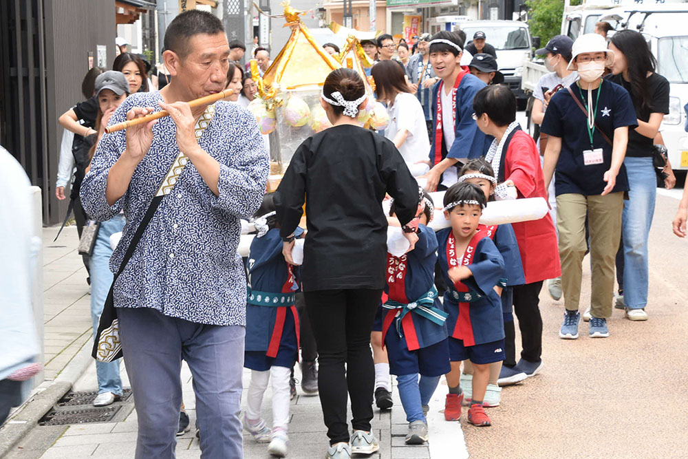 お囃子の後ろを神輿を担いで歩く幼稚園児の画像