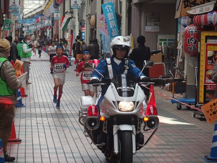 商店街を駆ける選手と先導白バイ